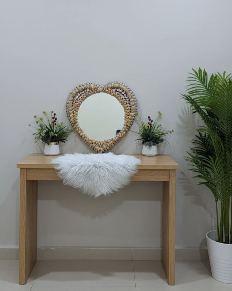 Wooden console table with a heart-shaped mirror, white fur throw, and potted plants against a plain wall.