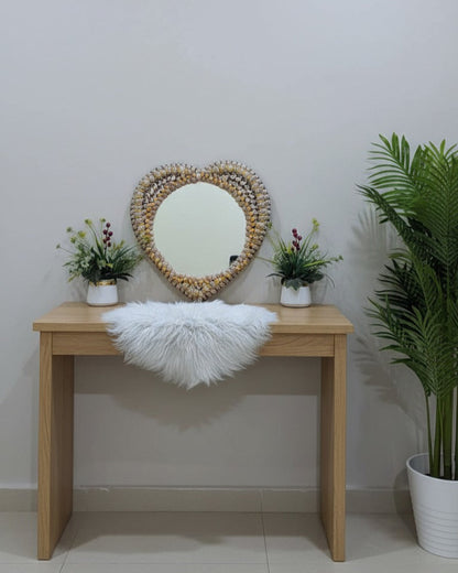 Wooden console table with a heart-shaped mirror, white fur throw, and potted plants against a plain wall.