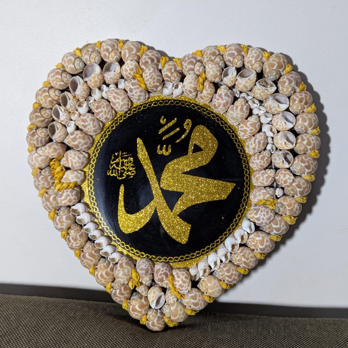 Heart-shaped decorative item made of shells with a central black circle featuring gold Arabic calligraphy on a white background.
