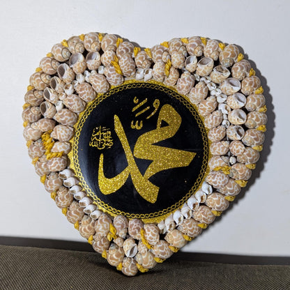 Heart-shaped decorative item made of shells with a central black circle featuring gold Arabic calligraphy on a white background.