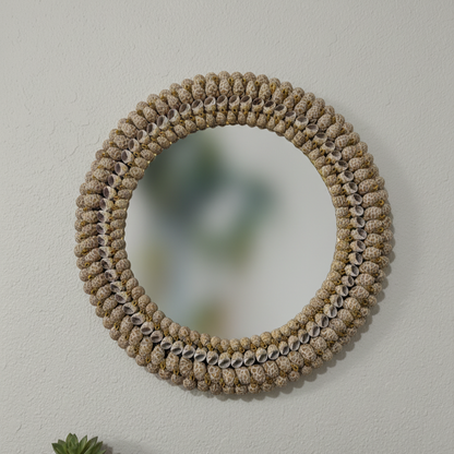 Round woven mirror on a plain wall with a wooden shelf below featuring a small plant and stones.